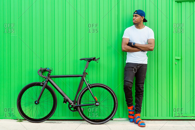 Young African American man in casual street style standing near bike against green wall on the street and looking away