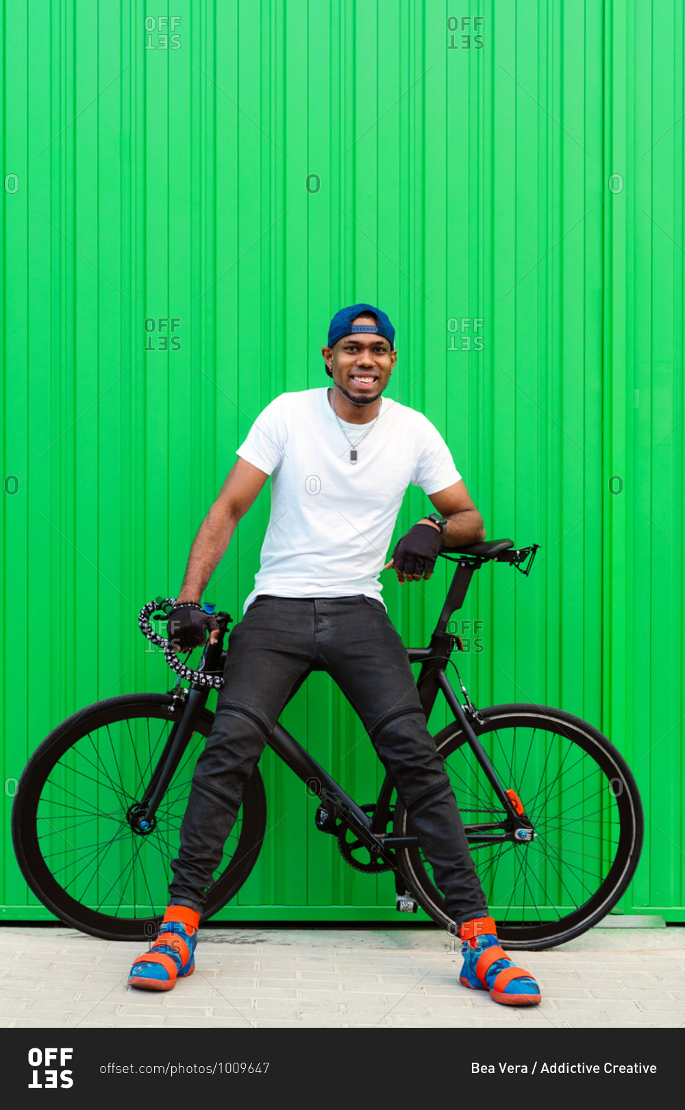 Full body of cheerful young African American man in casual street style wear and trendy shoes sitting on bike against green wall on street and looking away