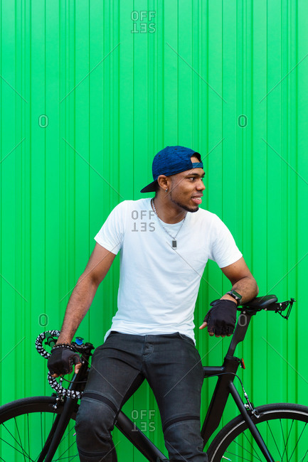 Happy young African American man in casual street style standing sitting on bike against green wall on street and looking away