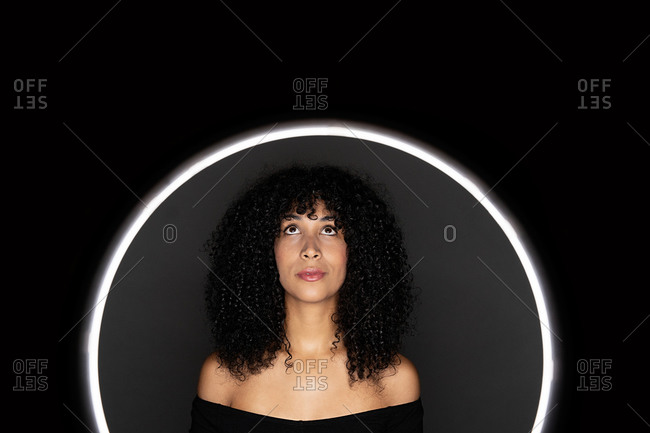 Thoughtful African American woman with curly hair standing in a light circle frame looking up in studio on black background