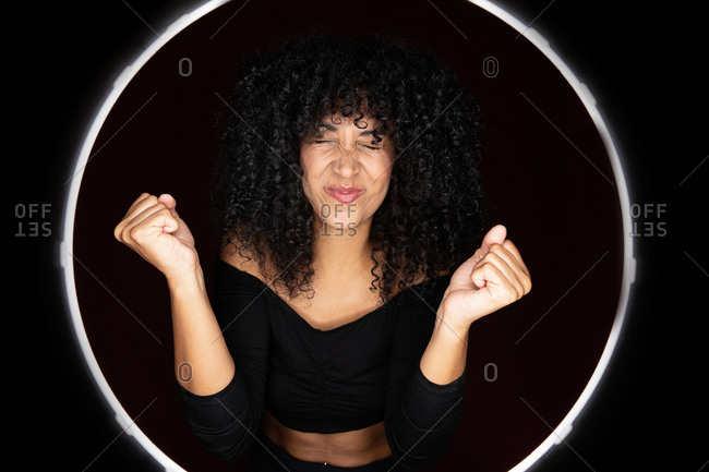 Cheerful excited African American woman with curly hair standing in a light circle frame with eyes closed in studio