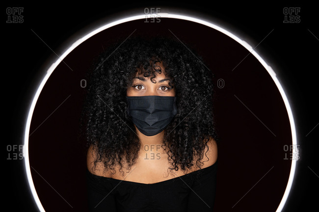 Thoughtful African American woman with curly hair wearing face medical mask against coronavirus standing in a light circle frame looking at camera in studio on black background