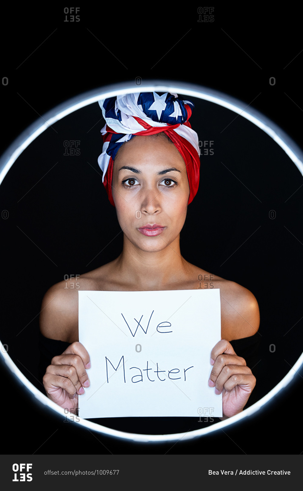 Portrait of a black female framed on a led ring wearing proudly the United States of America colors as a head wrap while holding a whiteboard with the peaceful message black lives matter looking at camera