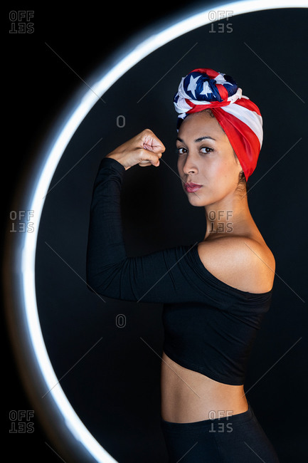 Side view of thoughtful and powerful black female standing in front of a led ring light wearing proudly the United States of America colors as a head wrap while showing arm muscle and strength