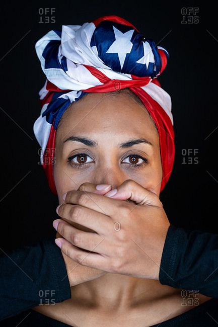 Portrait of scared black female wearing the United States of America colors as a head wrap while having her mouth covered with hands on black background