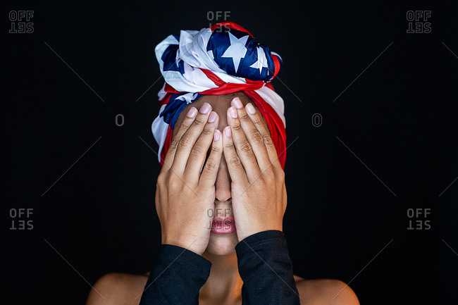 Portrait of unrecognizable scared black female wearing proudly the United States of America colors as a head wrap while covering eyes with hands on black background