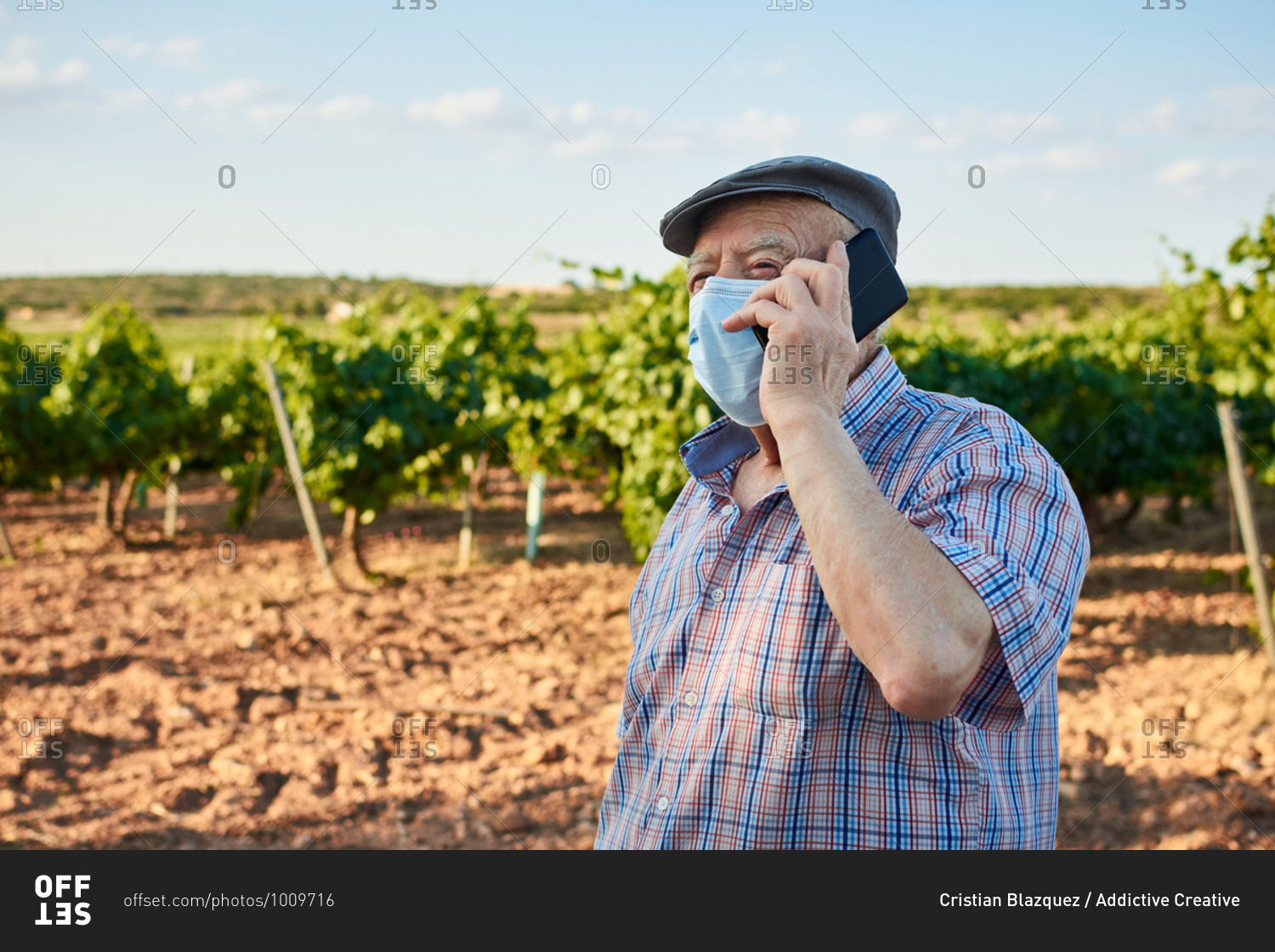 Senior male in protective mask standing on rural road and reading message on mobile phone in summer day