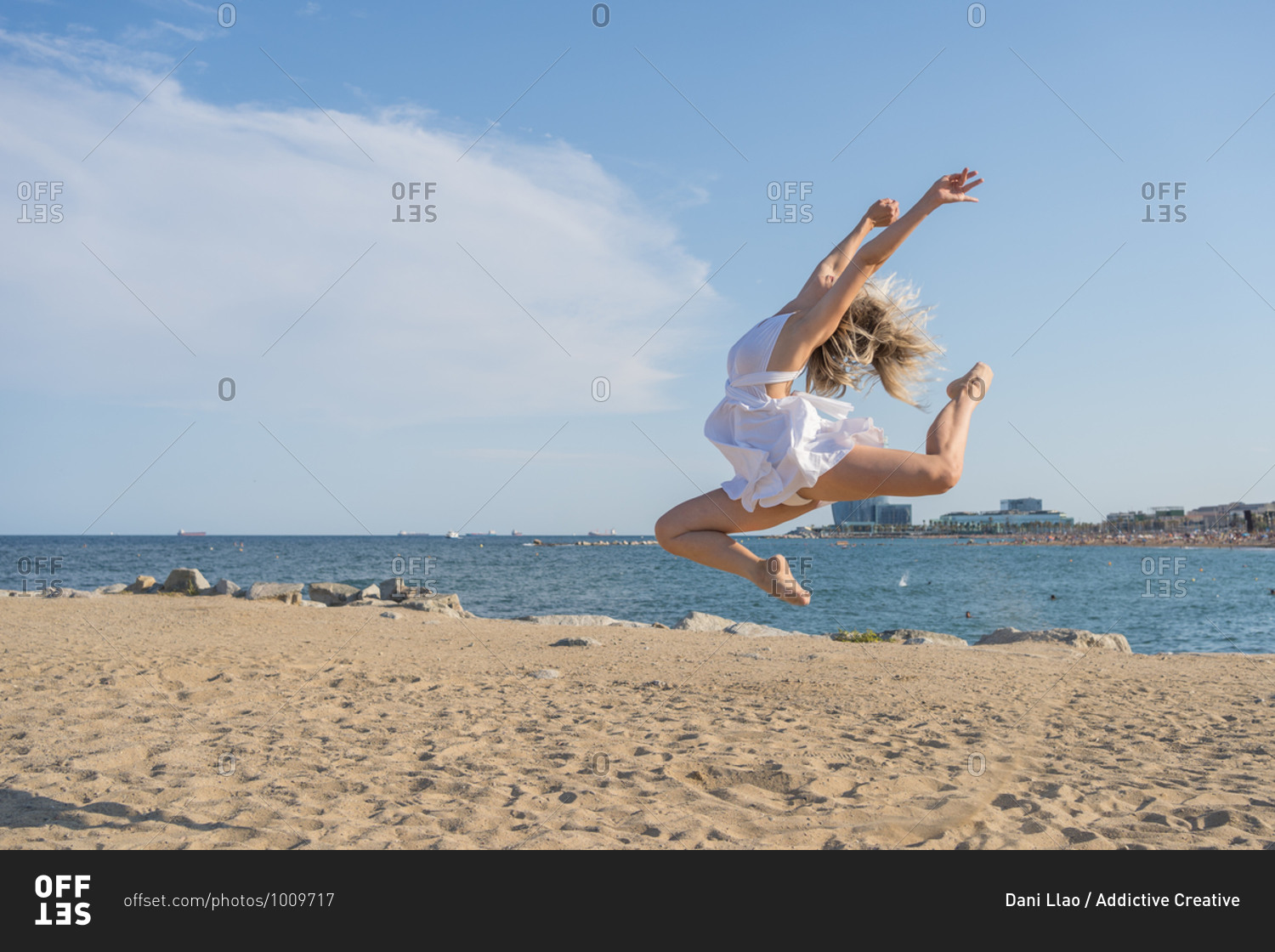Full length side view of unrecognizable barefoot female dancer in white dress jumping up while performing dynamic dance moves with arms raised on sandy beach