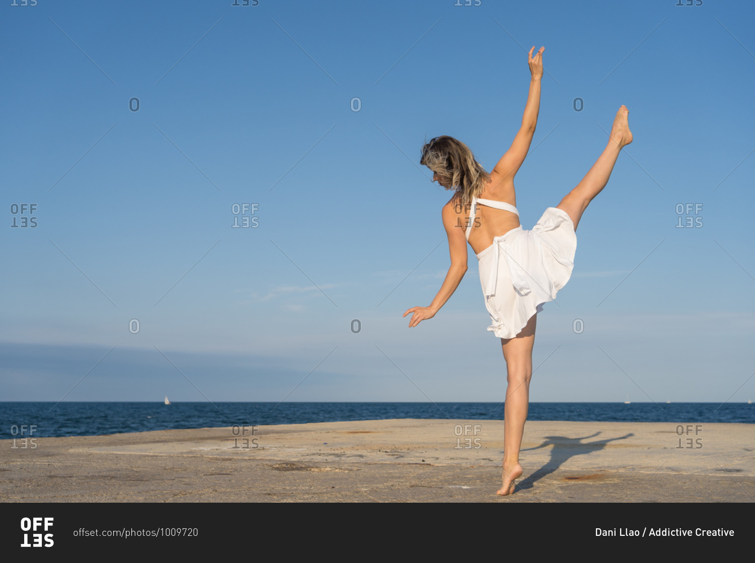 Full body barefoot female dancer in white dress standing on tiptoe and stretching leg while practicing ballet movements on sunny seaside