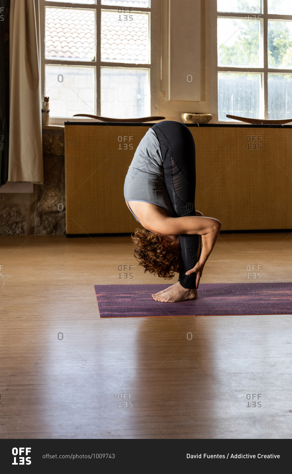 Full body side view of unrecognizable female in sportswear performing Standing Forward Bend pose during Shakti yoga session in light studio with wooden interior