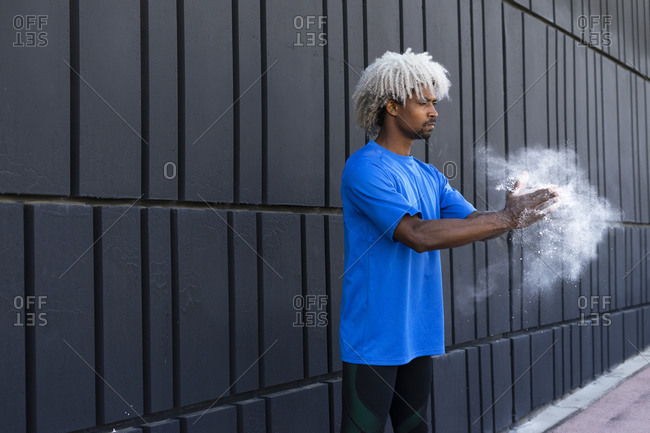 African American male athlete with dyed hair clapping hands with chalk powder while preparing for training near wall of modern building