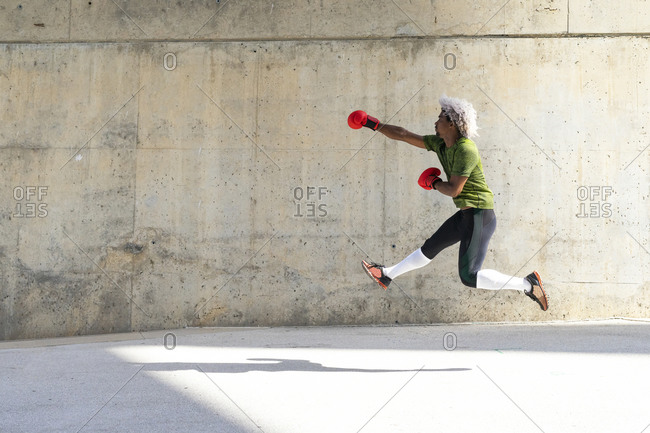 Side view of African American sportsman leaping and practicing jab against weathered concrete wall during boxing training on street