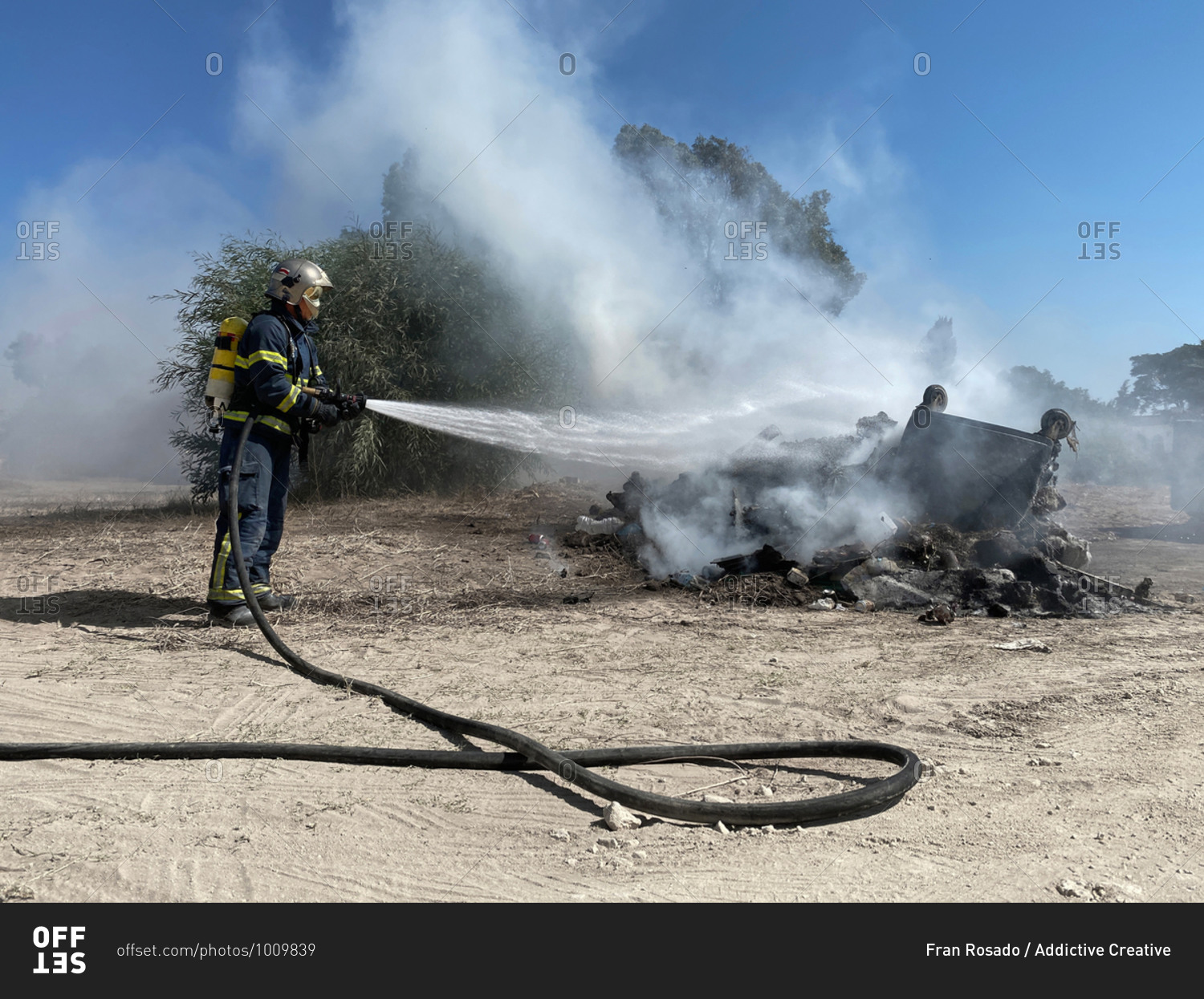 Side view of brave fireman in protective uniform standing with hose and extinguishing fire on dump in nature