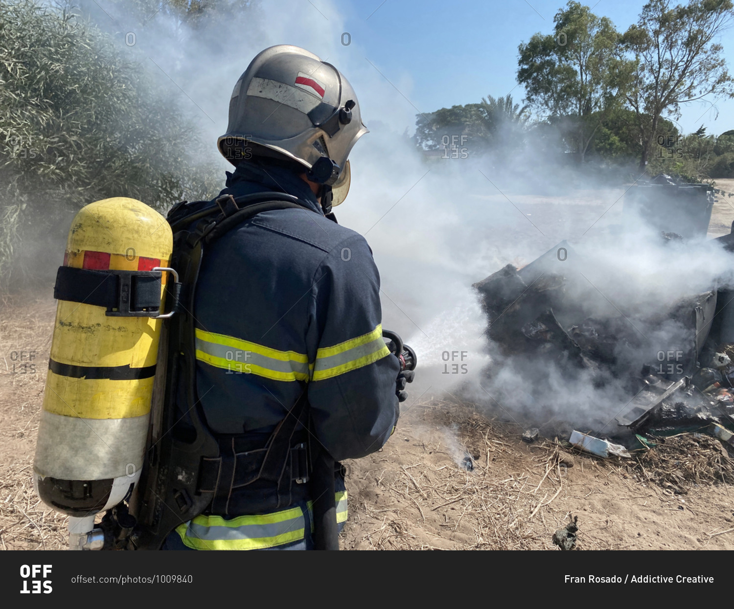 Back view of brave fireman in protective uniform standing with hose and extinguishing fire on dump in nature