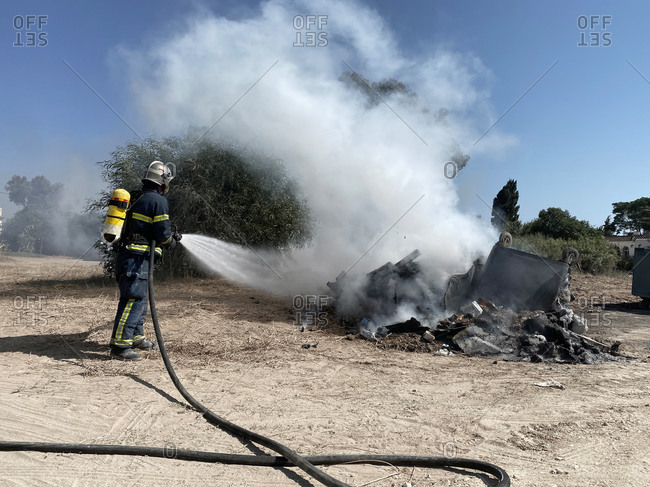 Back view of brave fireman in protective uniform standing with hose and extinguishing fire on dump in nature