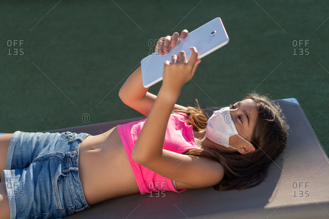 Smiling teenage girl lying on lounger in backyard and watching cartoon while using tablet and enjoying weekend in summer