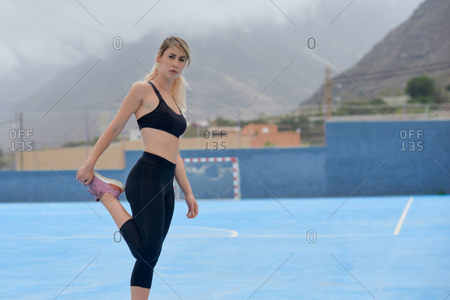 Full body side view of serious young female runner in black leggings and top stretching legs while standing near fence on sports ground and preparing for training