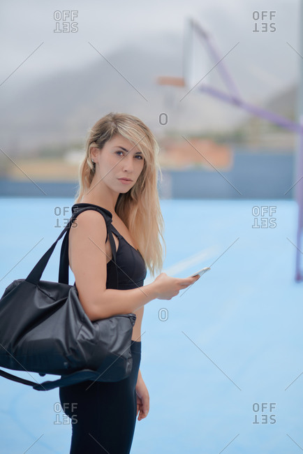 Full body side view of happy young slim sportswoman in black activewear with bag over shoulder standing on sports ground and messaging on mobile phone