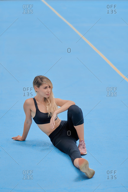 Full body of focused young fit female in sportswear sitting on blue ground and bending forward while stretching body during fitness workout on sports ground