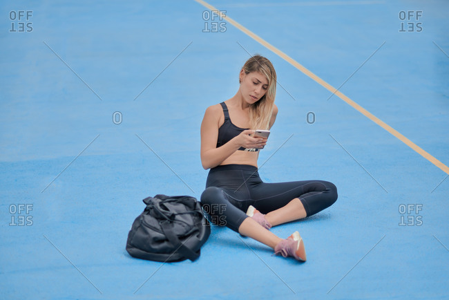 Full body young athletic female in sportswear with sports bag sitting on blue ground and browsing mobile phone while relaxing after training on sports ground