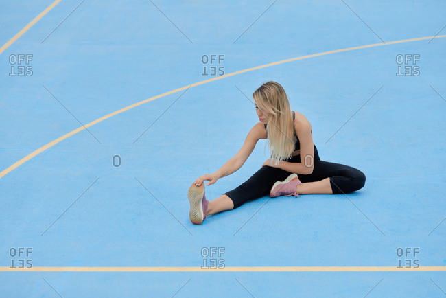 Full body of focused young fit female in sportswear sitting on blue ground and bending forward while stretching body during fitness workout on sports ground