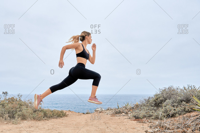 Low angle side view of young fit female in sports bra and leggings running fast along sandy seashore during fitness workout in summer day