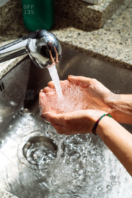 Unrecognizable person cleaning hands with water while standing near sink in bathroom lit by sunlight