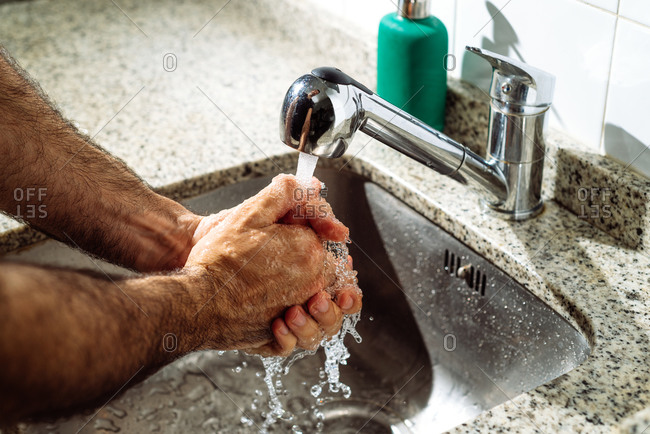 Unrecognizable person cleaning hands with water while standing near sink in bathroom lit by sunlight