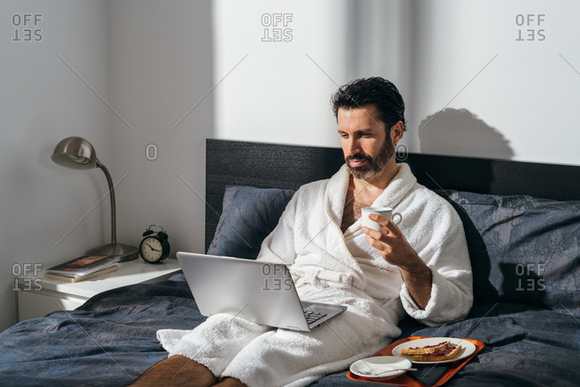 Handsome male freelancer in bathrobe sitting on bed with breakfast and cup of fresh coffee while working on laptop in morning