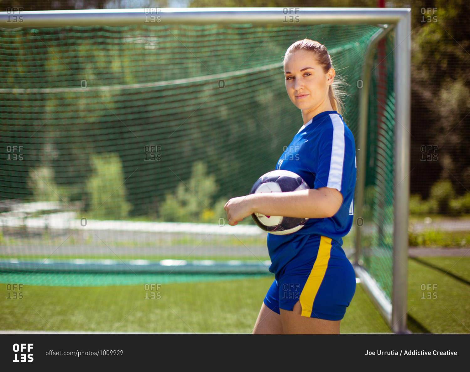 Side view of female football player with ball looking at camera while standing near goal during training on sunny day on field