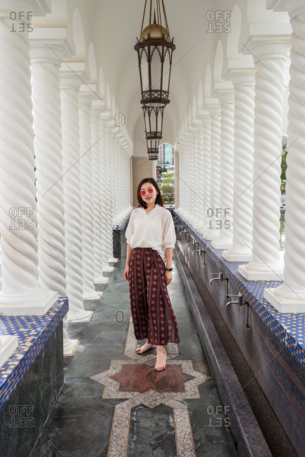 Full body of ethnic female traveler in summer outfit standing in ornamental walkway with stone columns of Omar Ali Saifuddien Mosque during holiday and looking at camera