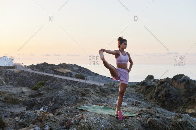 Fit young woman in her mountain one legged pose while practicing yoga on a rocky coast in the early morning