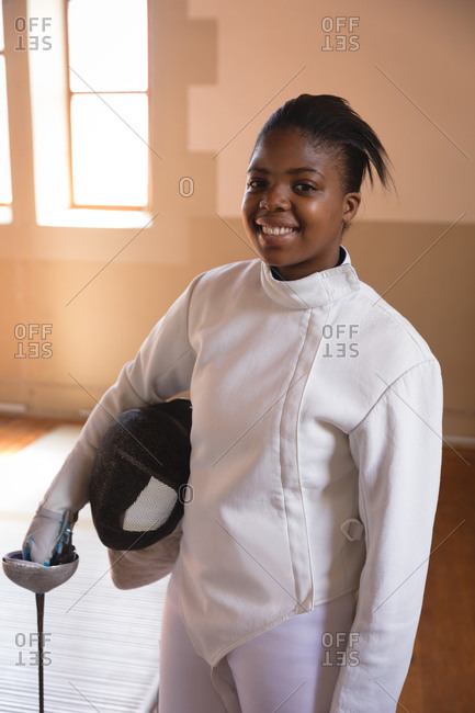 Portrait of an African American sportswoman wearing protective fencing outfit during a fencing training session, looking at camera and smiling, holding an epee and mask. Fencers training at a gym.