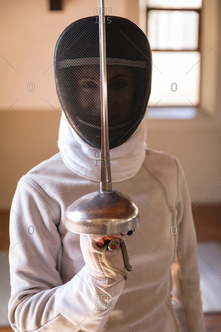 Portrait of Caucasian sportswoman wearing protective fencing outfit during a fencing training session, looking at camera and smiling, holding an epee in defensive position. Fencers training at a gym.