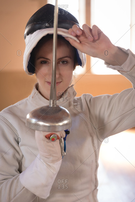Portrait of Caucasian sportswoman wearing protective fencing outfit during a fencing training session, looking at camera and smiling, holding an epee. Fencers training at a gym.
