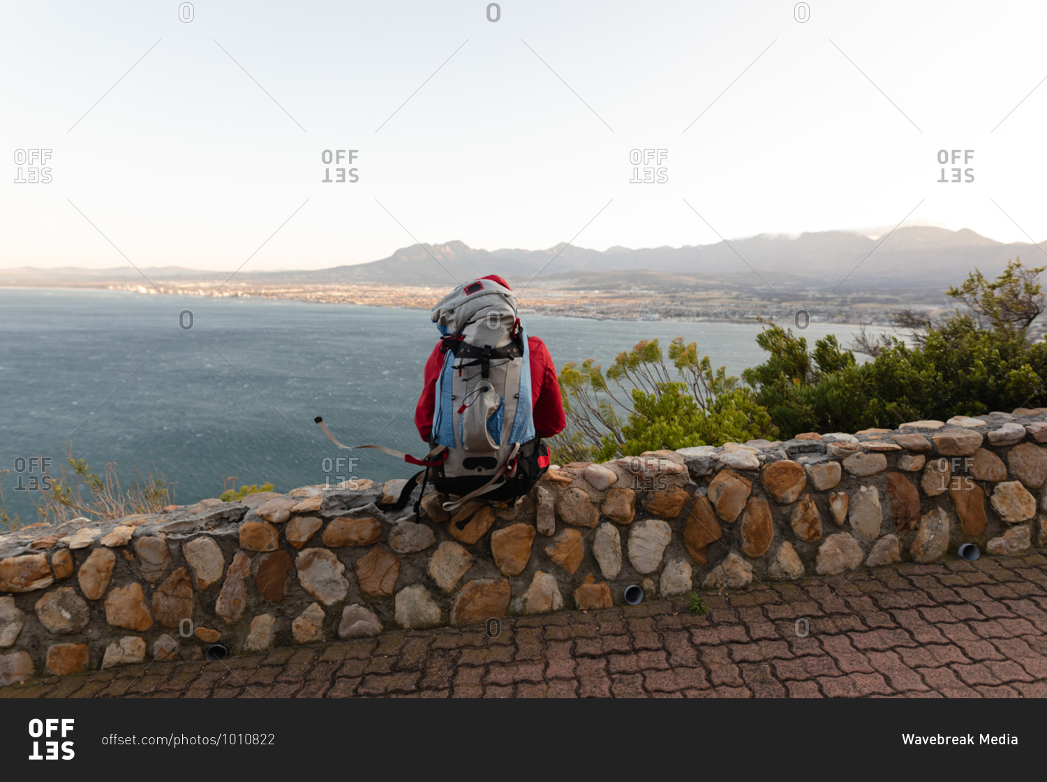 Rear view of a disabled mixed race male athlete with prosthetic leg, enjoying his time on a trip to the mountains, hiking, sitting on a wall on the road by the sea. Active lifestyle with disability.
