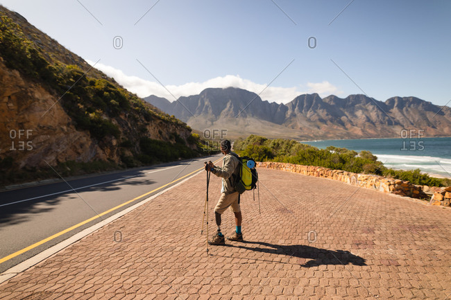 A fit, disabled mixed race male athlete with prosthetic leg, enjoying his time on a trip to the mountains, hiking with sticks, walking on the road by the sea. Active lifestyle with disability.