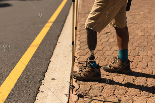 Low section of a fit, disabled mixed race male athlete with prosthetic leg, enjoying his time on a trip to the mountains, hiking with sticks, standing on the road. Active lifestyle with disability.
