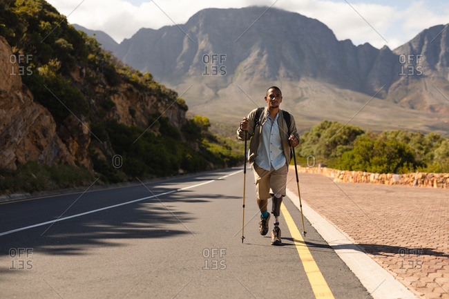 A fit, disabled mixed race male athlete with prosthetic leg, enjoying his time on a trip to the mountains, hiking with sticks, walking on the road in the mountains. Active lifestyle with disability.