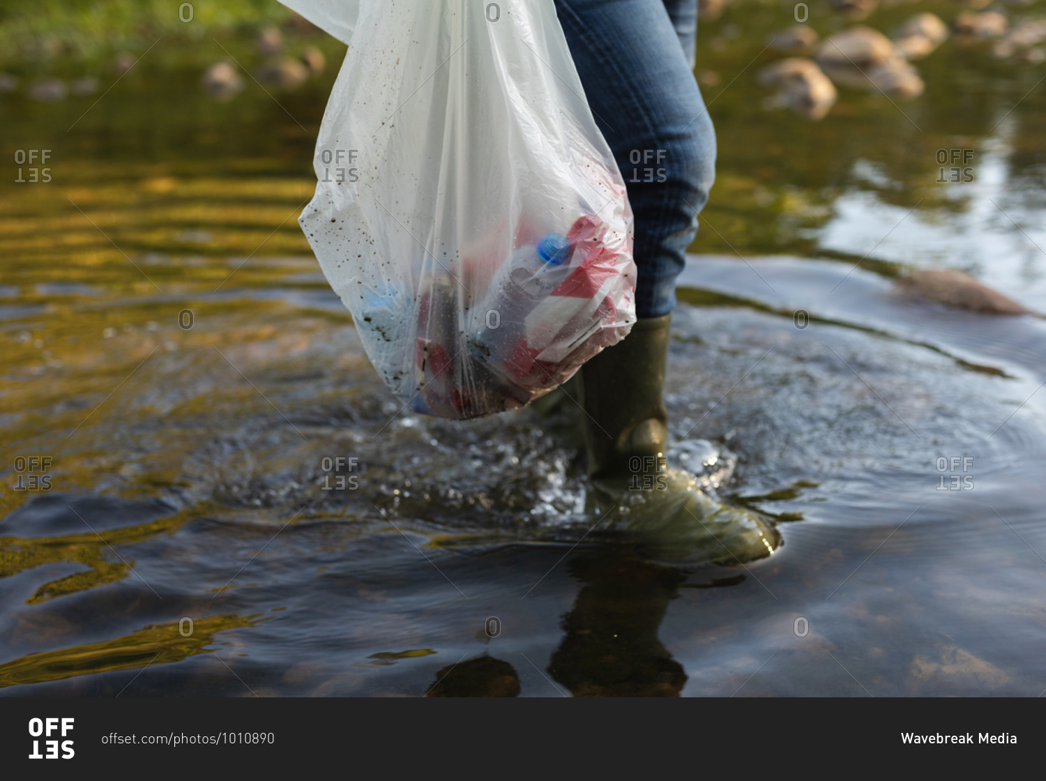 Low section of conservation volunteer cleaning up river in the countryside, carrying bag full of rubbish. Ecology and social responsibility in rural environment.