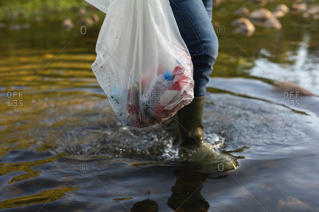 Low section of conservation volunteer cleaning up river in the countryside, carrying bag full of rubbish. Ecology and social responsibility in rural environment.
