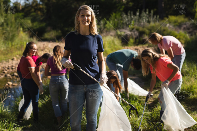 Portrait of Caucasian female conservation volunteer cleaning up river in the countryside, her friends picking up rubbish in the background. Ecology and social responsibility in rural environment.