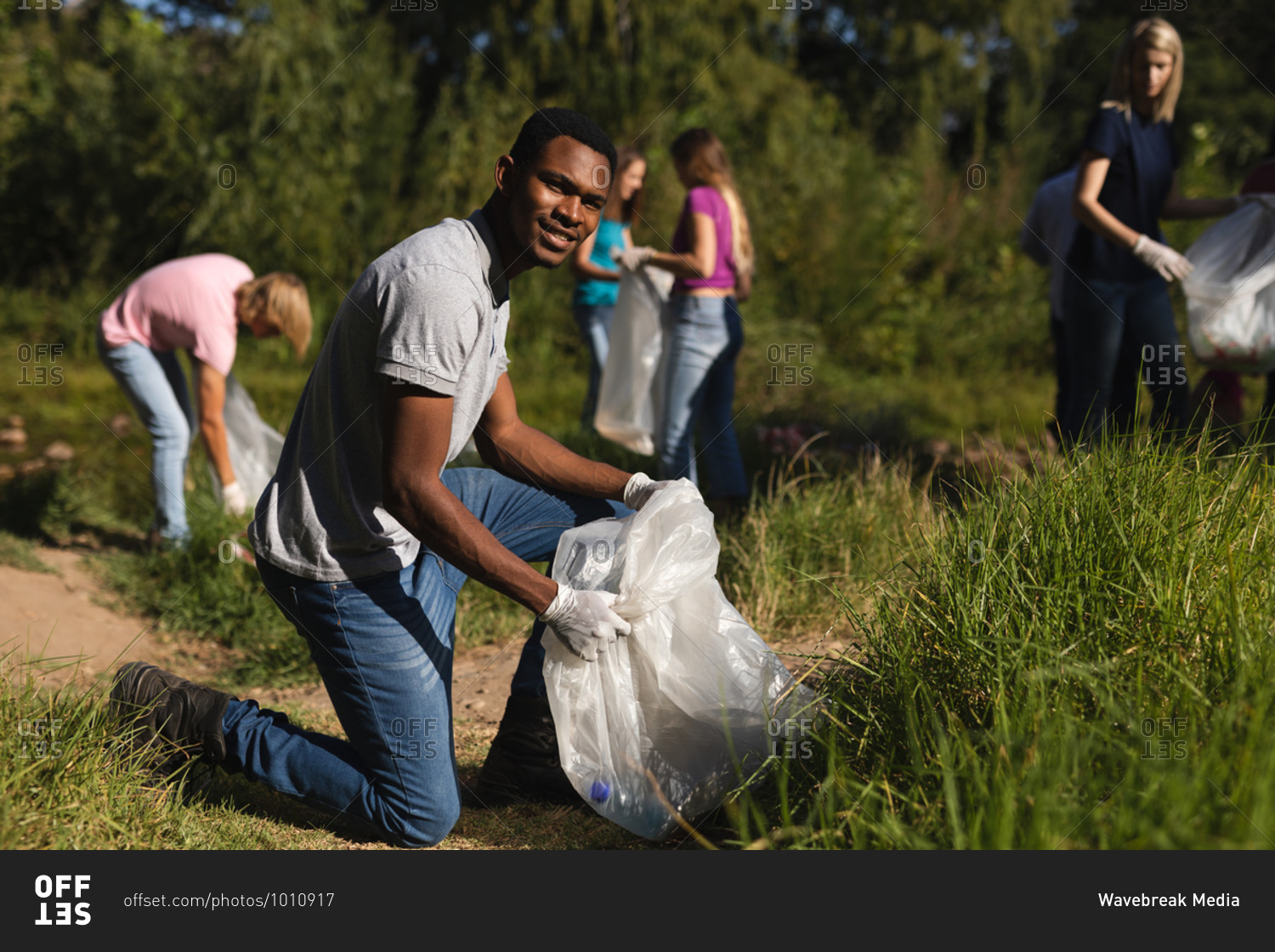 Portrait of African American male conservation volunteer cleaning up river in the countryside, his friends picking up rubbish in the background. Ecology and social responsibility in rural environment.