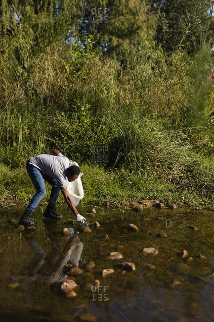 African American male conservation volunteer cleaning up river in the countryside, picking up rubbish. Ecology and social responsibility in rural environment.