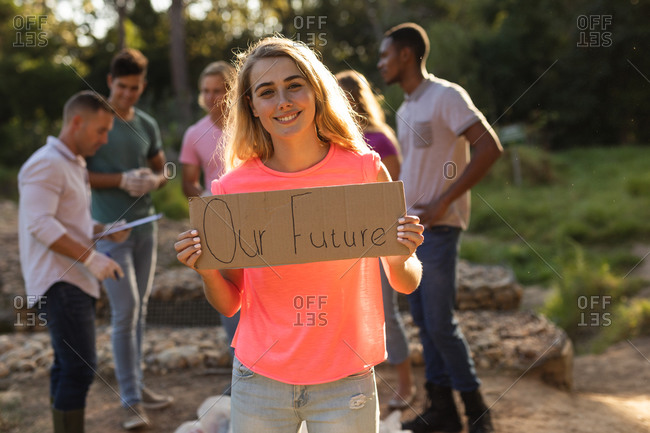 Portrait of happy Caucasian female conservation volunteer cleaning up the countryside with board Our Future, her friends in the background. Ecology and social responsibility in rural environment.