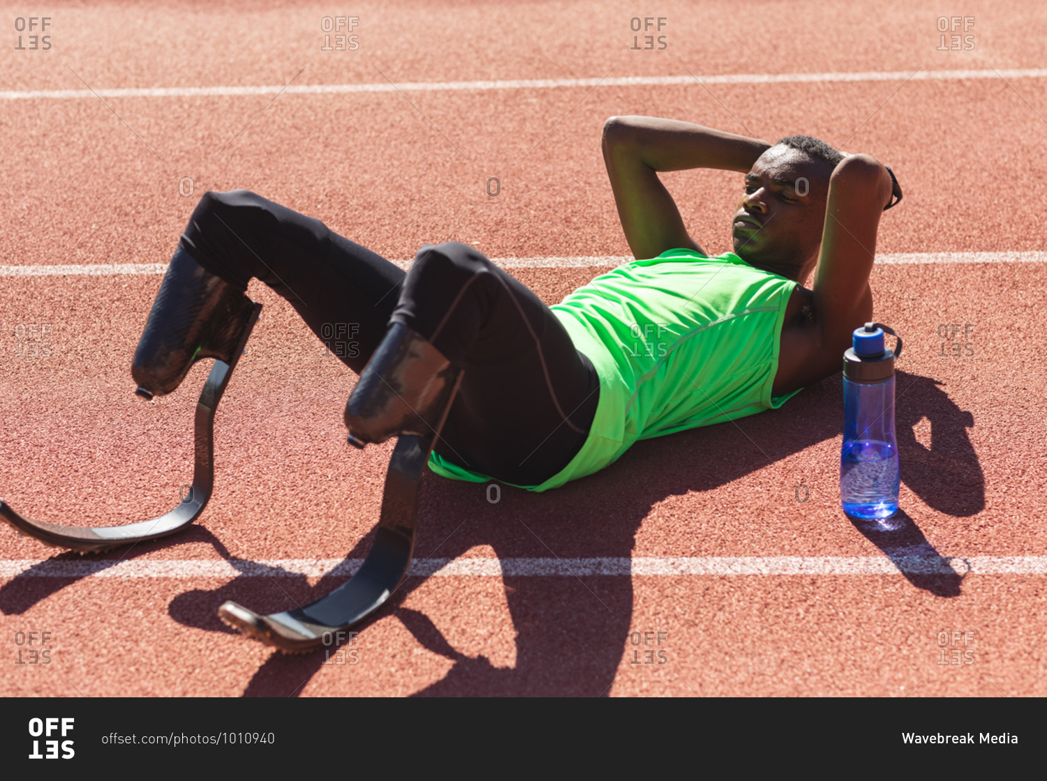 Fit, mixed race disabled male athlete at an outdoor sports stadium, lying on race track after race with water bottle wearing running blades. Disability athletics sport training.