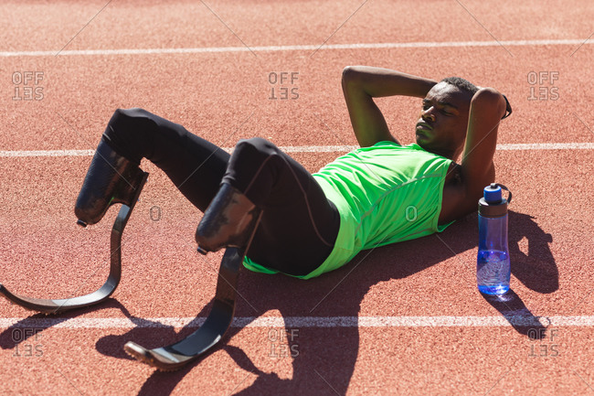 Fit, mixed race disabled male athlete at an outdoor sports stadium, lying on race track after race with water bottle wearing running blades. Disability athletics sport training.