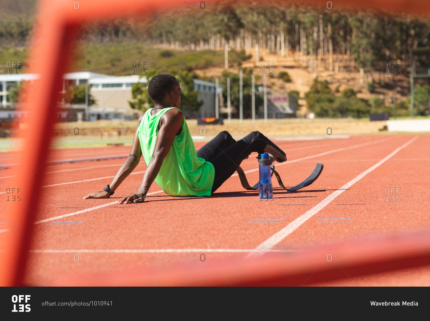 Fit, mixed race disabled male athlete at an outdoor sports stadium, sitting on race track after race with water bottle wearing running blades. Disability athletics sport training.