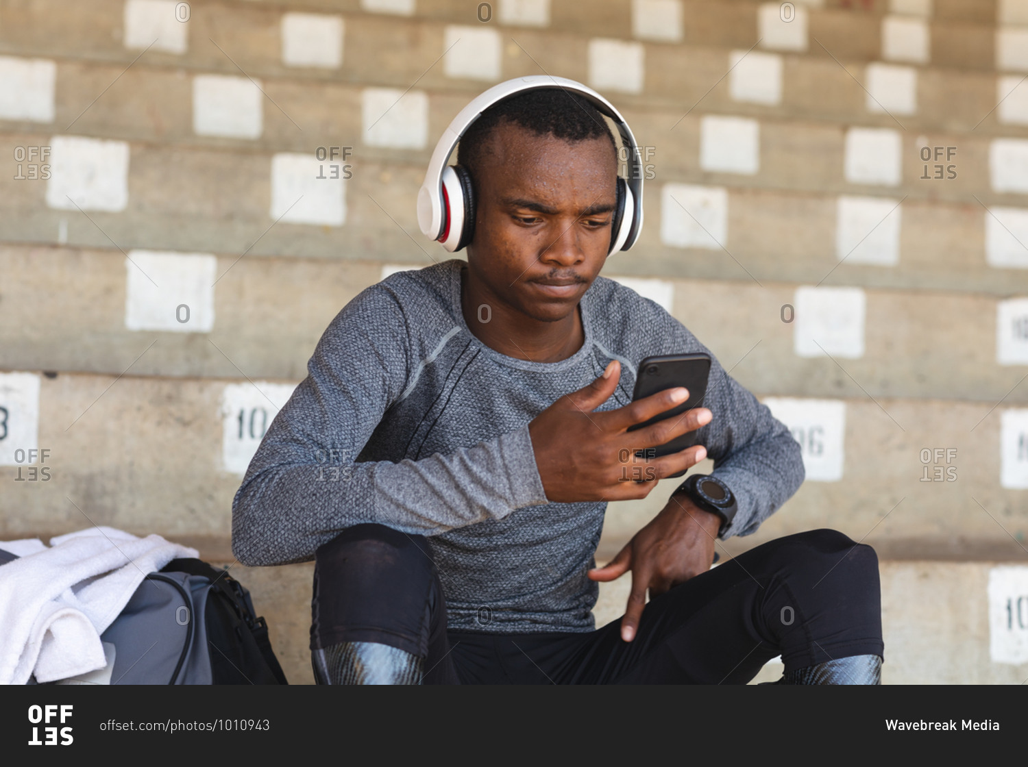 Fit, mixed race disabled male athlete at an outdoor sports stadium, sitting in the stands wearing headphones using smartphone wearing running blades. Disability athletics sport training.