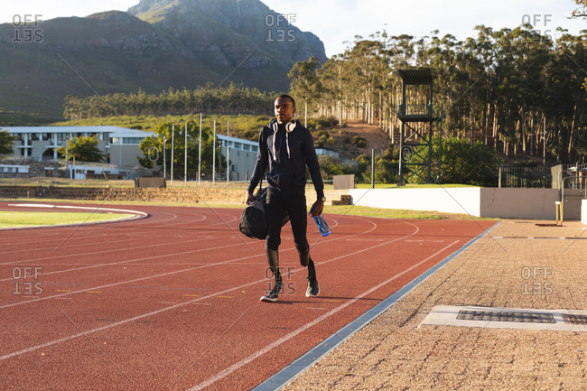 Fit, mixed race disabled male athlete at an outdoor sports stadium, walking with gym bag and water bottle on race track wearing running blades. Disability athletics sport training.